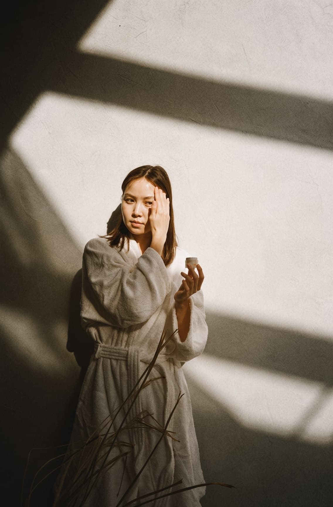Woman in a bathrobe applying moisturizer from a jar in warm indoor light