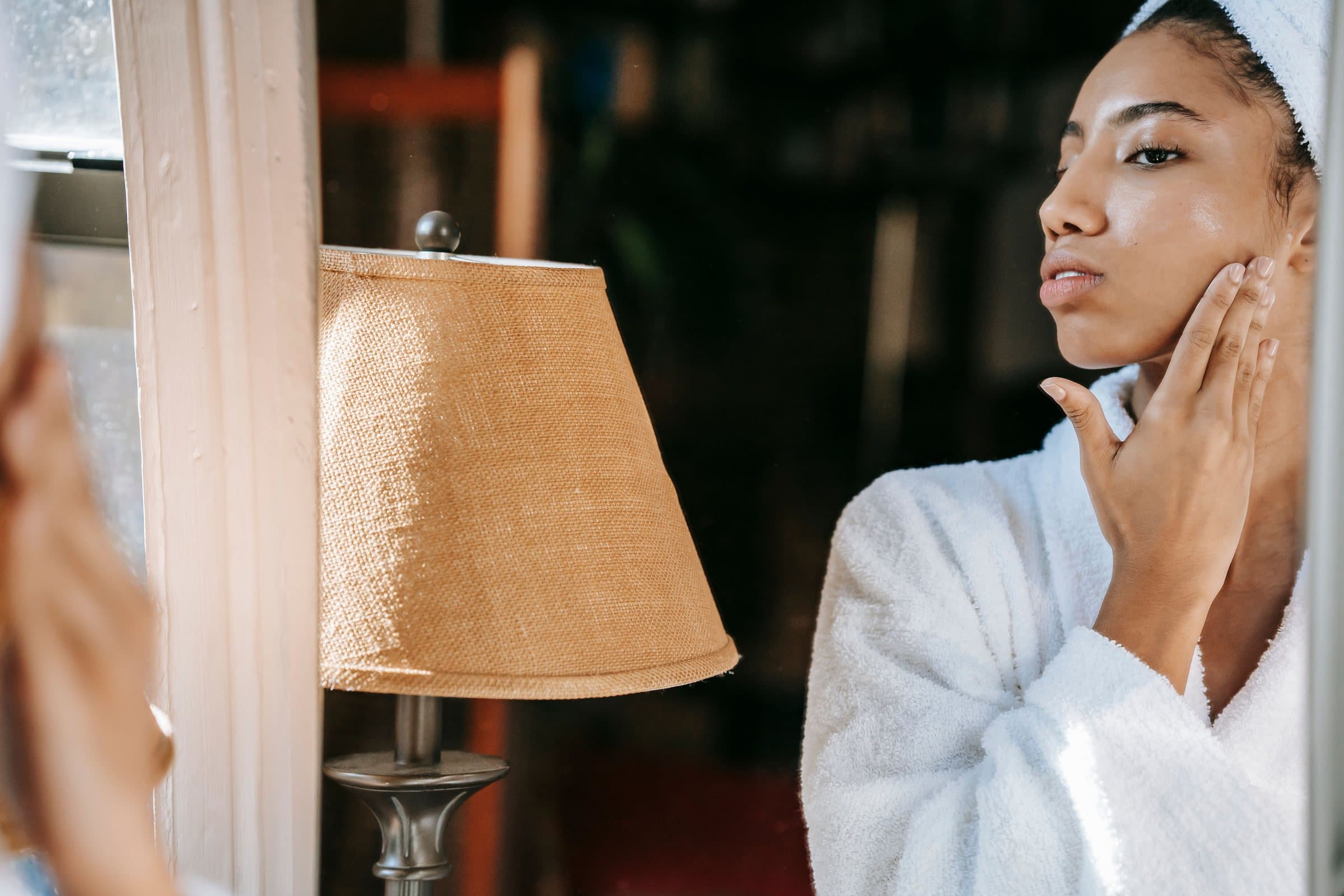 Woman in a white bathrobe and hair towel gently touching her cheek at a mirror in warm lamplight